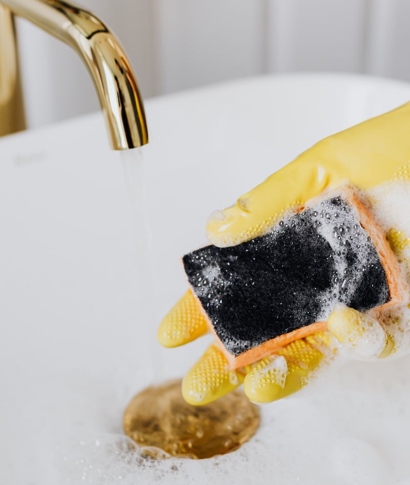Eine Hand in einem gelben Reinigungshandschuh hält einen schwarzen Schwamm unter fließendes Wasser aus einem goldenen Wasserhahn, wahrscheinlich im Begriff, Reinigungsarbeiten zu beginnen oder einen Reinigungsvorgang durchzuführen.