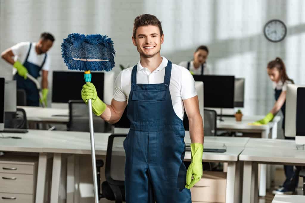 Lächelnder Reinigungstechniker in Arbeitskleidung und Handschuhen hält einen Wischmopp, mit Kollegen im Hintergrund, die ein Büro reinigen.